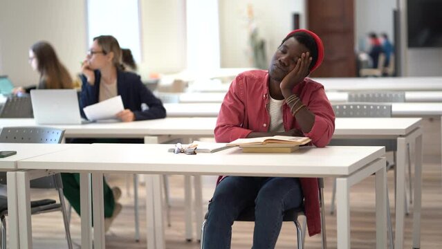 Sleepy lazy African man falls asleep sits in public library tired after long time reading literature. Exhausted black guy dozing with head resting on desktop with books on university campus