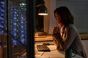 Asian young businesswoman sitting at her workplace in front of computer and working overtime in...