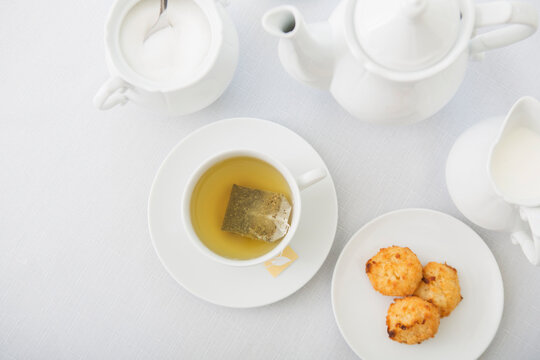 Cup of tea in porcelain white teacup with saucer, sugar bowl, creamer, teapot and plate of coconut macaroons, studio shot