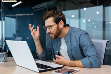 Dissatisfied and angry businessman looking at laptop screen, man in casual shirt working inside...