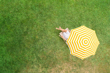 Woman in a white dress sitting on deck chair under yellow umbrella  on the green grass sunbathes at summer day. Top view, drone, aerial view. - 555505665