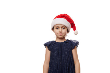Isolated portrait on white background of a Caucasian 5-6 years old child, cute little girl in elegant navy dress and Santa hat, smiles looking confidently at camera. Christmas and New Year's concept