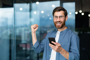 Portrait of a modern businessman in the middle of the office, the man is looking at the camera with a smartphone and celebrating the victory, the man is holding the phone in his hands and raising his