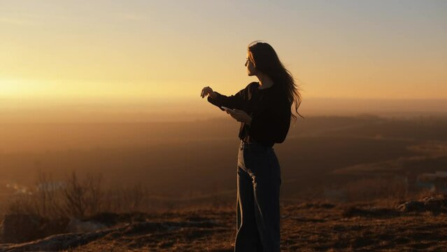A young slim girl woman stands on a hill at sunset with a beautiful view, holds a smartphone in her hands, looks at the sunset and covers the sunlight with her palm