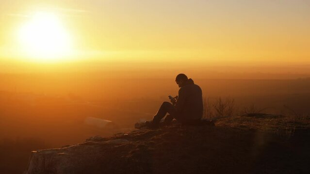 Silhouette of a man sitting on top of a mountain rock with an incredible view at sunset with a smartphone in his hands