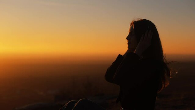 Silhouette of happy smiling woman girl against yellow sky at evening sunset, sitting on top of mountain with incredible view, wearing headphones and listening to music, traveler, outdoor walk