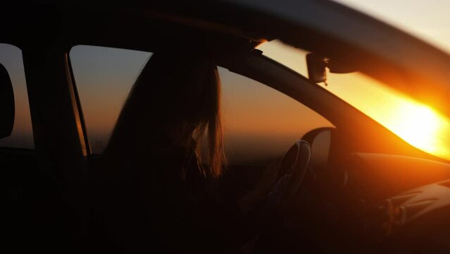 Side view, silhouette of a happy young woman driving a car and dancing to music while driving at sunset. Beautiful woman listening to energetic music and smiling. The concept of joy and happiness