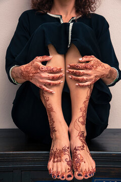 Low section of woman sitting indoors, showing legs, feet and hands painted with henna in arabic style, wearing a typical black, arabic, muslim dress, studio shot
