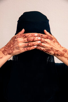 Close-up portrait of woman wearing black muslim hijab and muslim dress, hands covering eyes and showing hands painted with henna in arabic style, studio shot on whtie background