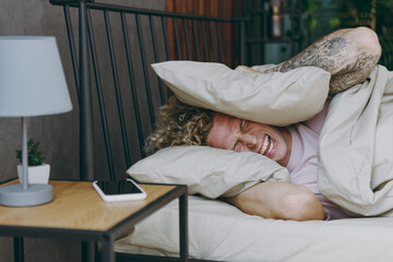 Young sad unhappy man in white t-shirt pajama he lying in bed cover ears with pillow from...