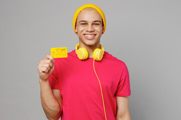 Young smiling happy fun cheerful man of African American ethnicity 20s he wearing pink t-shirt yellow hat headphones hold in hand mock up of credit bank card isolated on plain grey background studio.