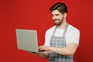 Young happy male housewife housekeeper chef cook baker IT man wear grey apron hold use work on laptop pc computer look for recipe isolated on plain red color background studio. Cooking food concept.