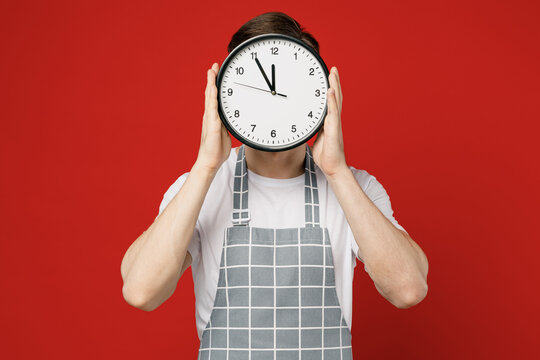 Young caucasian male housewife housekeeper chef cook baker man wears grey apron hold in hand cover face with clock show time for recipe isolated on plain red background studio. Cooking food concept.