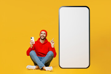 Full body young man wears red hoody hat near big huge blank screen point finger on mobile cell phone with workspace copy space mockup area smartphone chat online isolated on plain yellow background.
