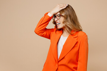 Young employee business woman corporate lawyer in classic formal orange suit glasses work in office put hand on head facepalm epic fail mistaken omg gesture isolated on plain beige background studio