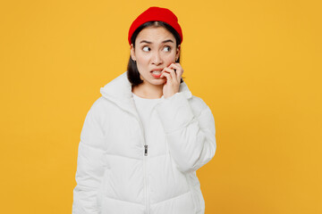 Young mistaken sad woman of Asian ethnicity wear white padded windbreaker jacket red hat look aside biting nails fingers isolated on plain yellow background studio portrait. People lifestyle concept.