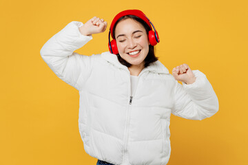 Young satisfied woman of Asian ethnicity wear white padded windbreaker jacket red hat headphones listen to music dance isolated on plain yellow background studio portrait. People lifestyle concept.