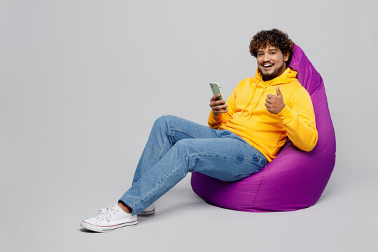 Full body young Indian man he wear casual yellow hoody sit in bag chair hold in hand use mobile cell phone show thumb up isolated on plain grey background studio portrait. People lifestyle portrait