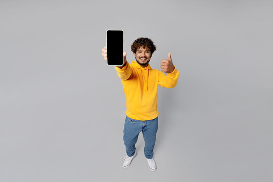 Full Body Smiling Overjoyed Happy Fun Cheerful Young Indian Man 20s He Wearing Casual Yellow Hoody Hold In Hand Use Mobile Cell Phone Show Thumb Up Isolated On Plain Grey Background Studio Portrait.