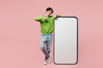 Full body happy young man of Asian ethnicity wear green hoody point index finger on big huge blank screen mobile cell phone smartphone with area isolated on plain pastel light pink background studio.