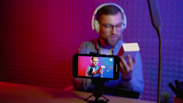 Excited overjoyed young male blogger unpacking boxe with new smartphone and doing live streaming. Attractive man using smart phone camera for creating content