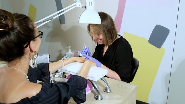 Young woman in glasses undergoes manicure sitting in modern beauty parlor. Smiling manicurist files shape of nails holding hands of client on soft stand
