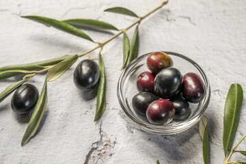 Black olives in glass bowl with black olives around and olive tree leaves, close up, top view