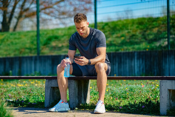 Obraz premium Resting after fitness. Young sportsman sitting on bench and checking his phone after exercising outdoor.