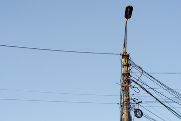 A concrete electric pole with a street lighting lamp at the top and numerous wires and coils on it against the blue sky