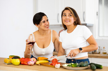 Two happy female friends preparing salad and drinking red wine together in modern kitchen