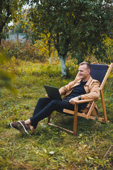 Fototapeta premium A young man works on a laptop outdoors. A young freelancer rests in the forest. Remote work, active recreation in the summer. Tourism, people concept - man sitting on a chair outdoors.