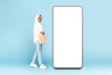 Happy black islamic woman shopaholic with shopping bag walking near big smartphone with empty screen, mockup, copy space