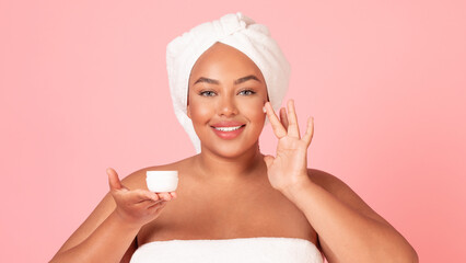 Beautiful african american oversize woman applying facial cream, holding jar in hand and smiling, pink background