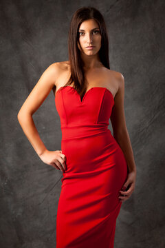 Portrait of young woman in red dress, standing and posing with hand on hip, looking at camera, grey background