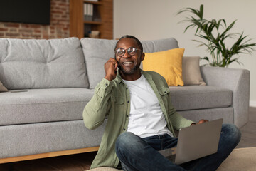 Glad smiling adult black businessman in glasses uses computer, calls by phone, gesticulates, sits...