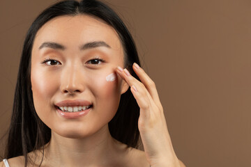 Portrait of asian lady applying anti-aging or moisturising face cream, posing over brown studio background, copy space