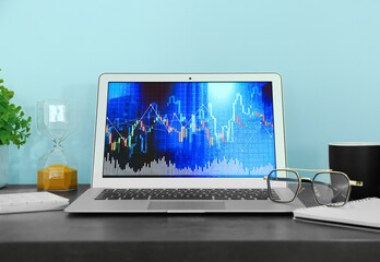 Laptop with stock data, hourglass and eyeglasses on table near blue wall. Finance trading