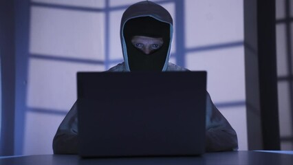 Masked hacker working in dark room, cracking password, hacking websites.
Hacker man cracking password from laptop in dark room doing cyber attack. Threatening. Illegal.
