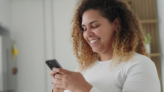 Happy black woman holding cell phone using smartphone device at home. Smiling young latin woman blogger subscribing new social media, buying in internet, ordering products online in apps.
