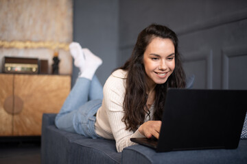 Happy caucasian woman using laptop pc computer, working or studying online, lying on sofa at home, free space