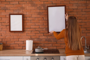 Young woman hanging blank frame on brick wall in kitchen, back view
