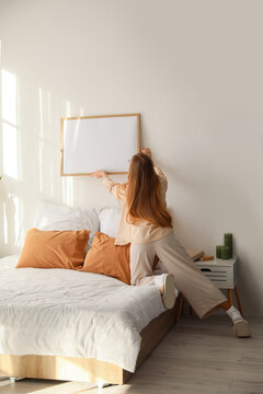 Young woman hanging blank frame on light wall in bedroom, back view