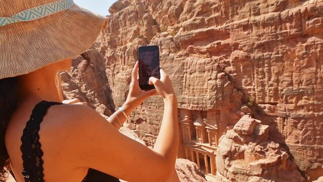 Gorgeous caucasian woman tourist sit on viewpoint in Petra ancient city over Treasury or Al-khazneh take smartphone photo. Jordan, one of seven wonders. UNESCO World Heritage site.