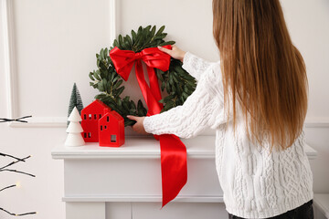 Young woman putting Christmas wreath on fireplace in living room