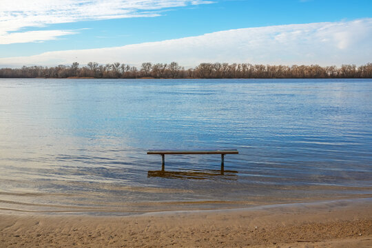 The water level in the river has risen. A wooden bench stands in the water. Forest on the horizon © Vlad Kazhan
