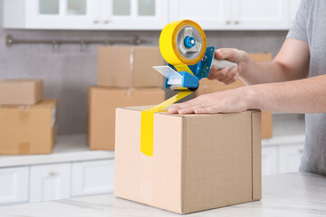 Man taping box with adhesive tape dispenser in kitchen, closeup
