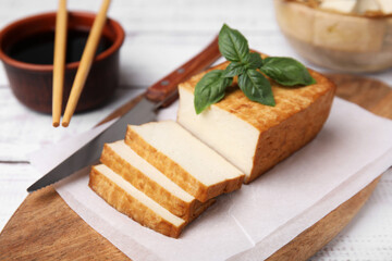 Board with smoked tofu, knife, basil and soy sauce on white wooden table