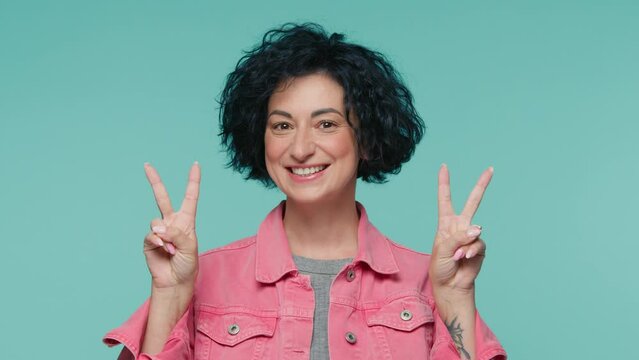 Gorgeous mature smiling woman with short dark curly hair showing V victory sign with fingers on both hands. Beautiful female 40s showing winning gesture slow motion people on blue studio background 4K