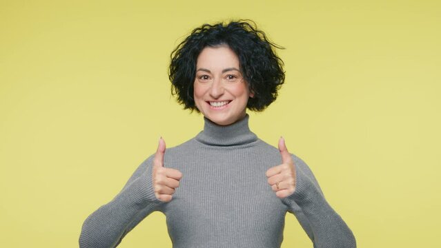 Cheerful Charming beautiful positive mature woman 40s showing thumbs up, looking happy at camera and enjoying posing on yellow studio background. Positive feedback, body language sign slow motion 4K