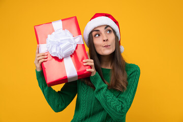 Studio shot of cute young woman wearing santa hat and green cozy knitted sweater posing over yellow background with present box in hands